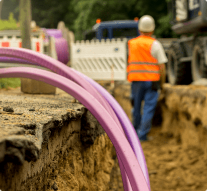 A man in an orange vest stands beside a purple pipe.