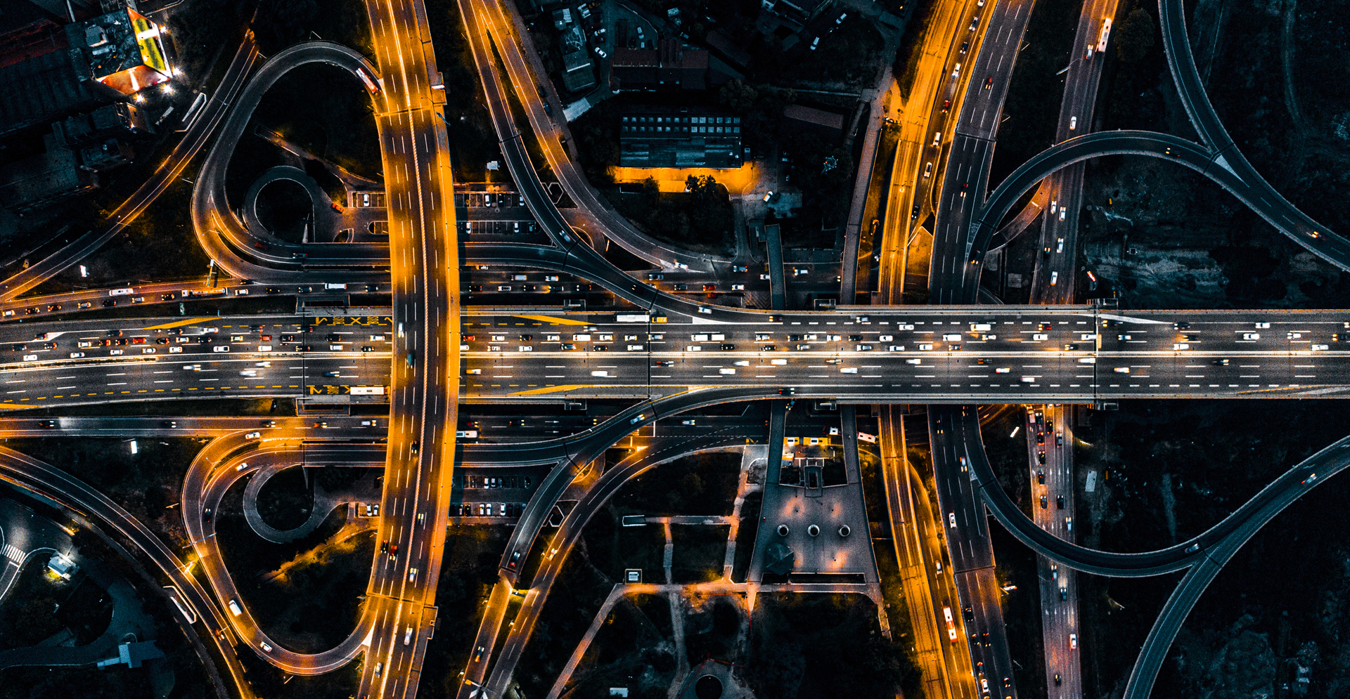 Aerial nighttime view of a large highway interchange with multiple overpasses and looping ramps illuminated by bright orange streetlights, with heavy traffic flowing in several directions.